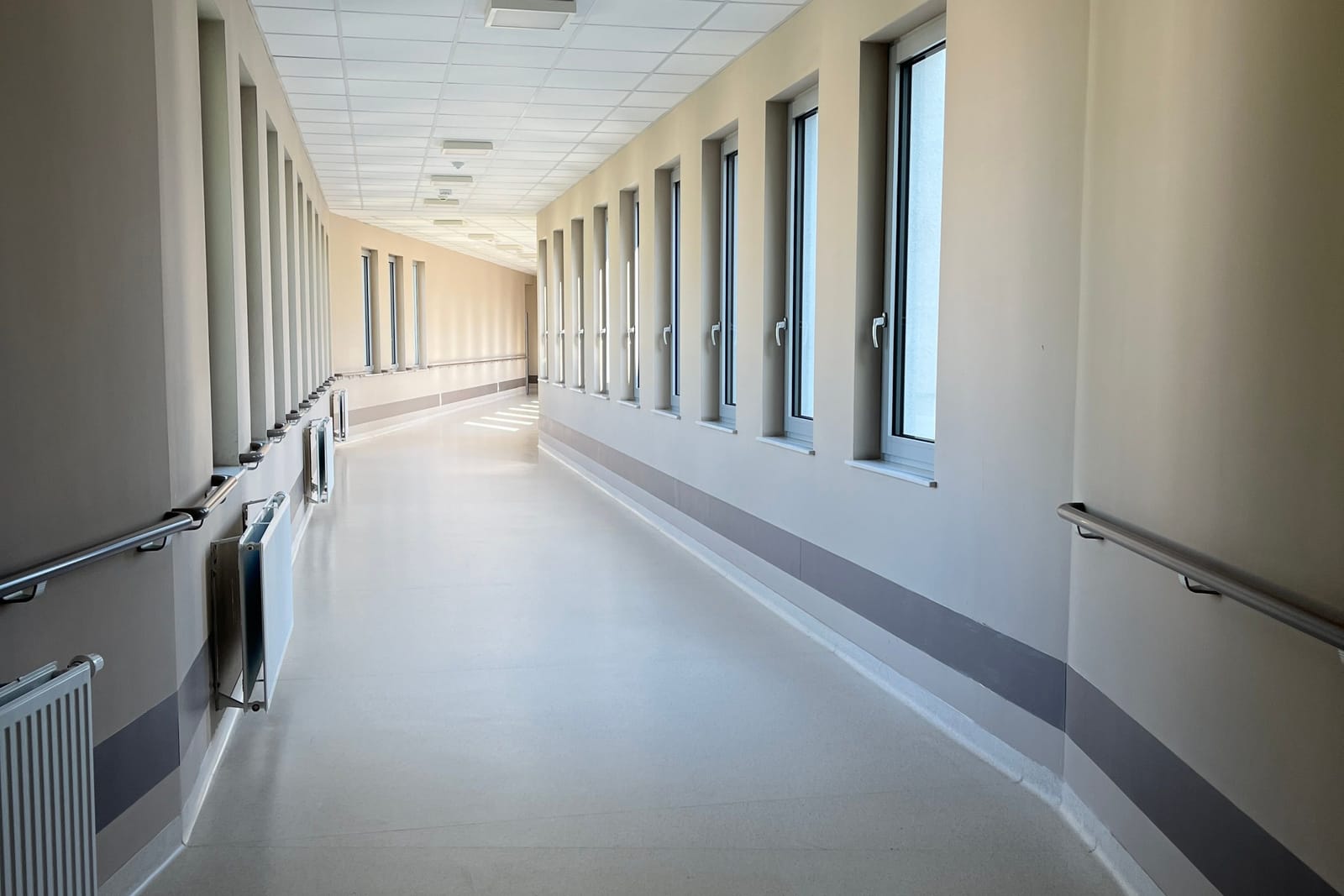 An empty, daylit hospital corridor with handrails and a row of tall windows