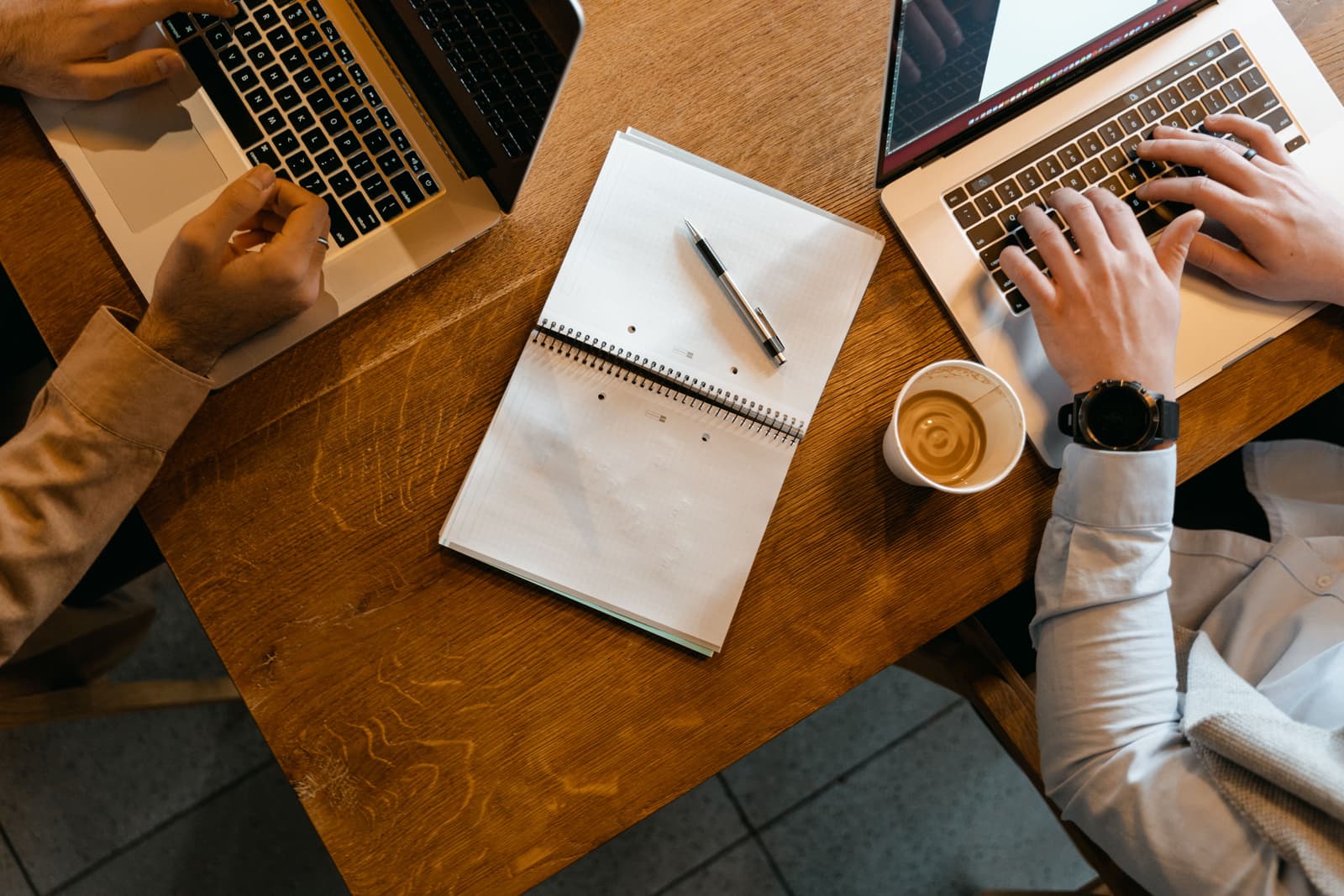 Seen from above, two people working on laptops across a wooden table, with a notebook and coffee between them