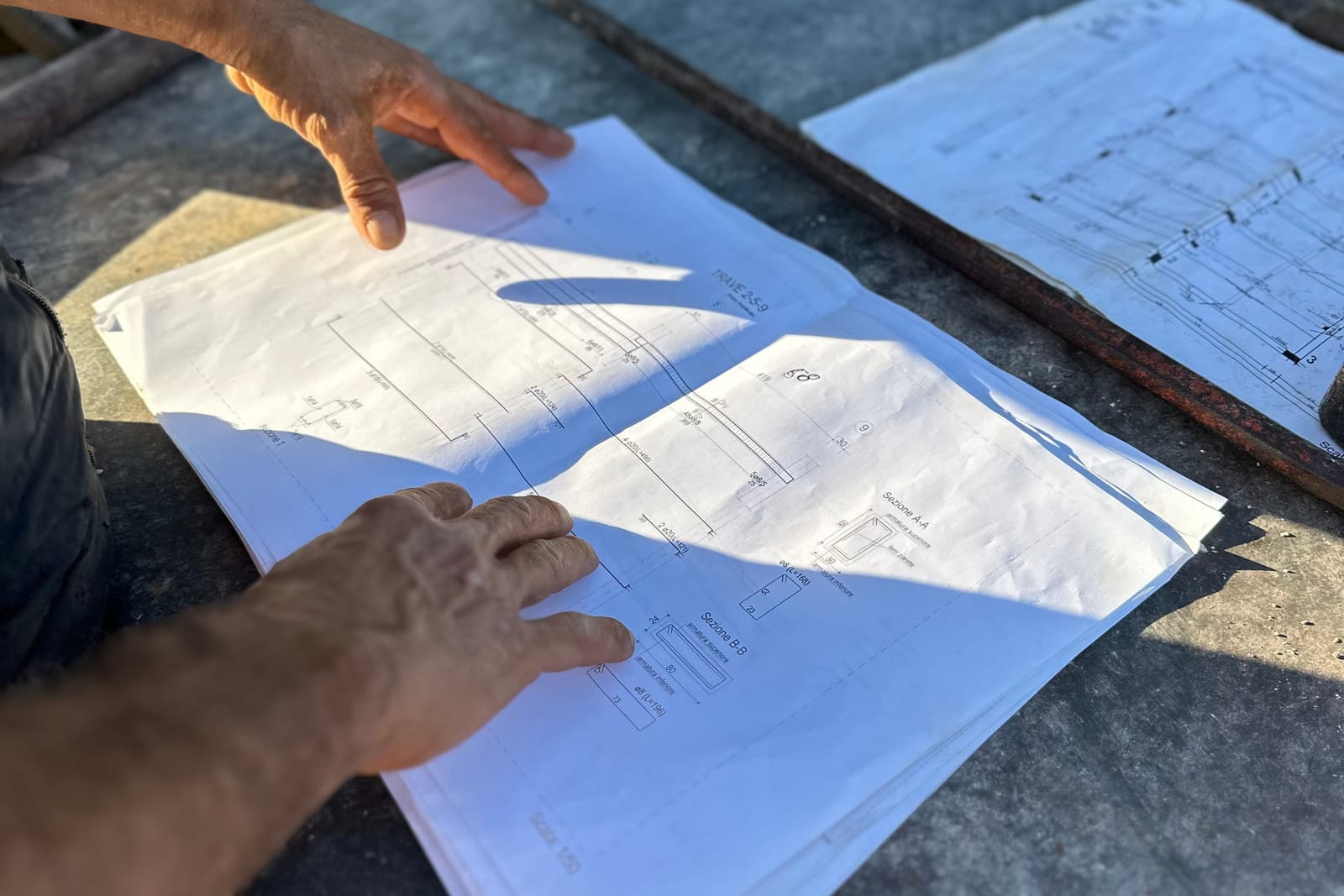 A pair of hands going over architectural drawings laid out on a bench in strong sunlight