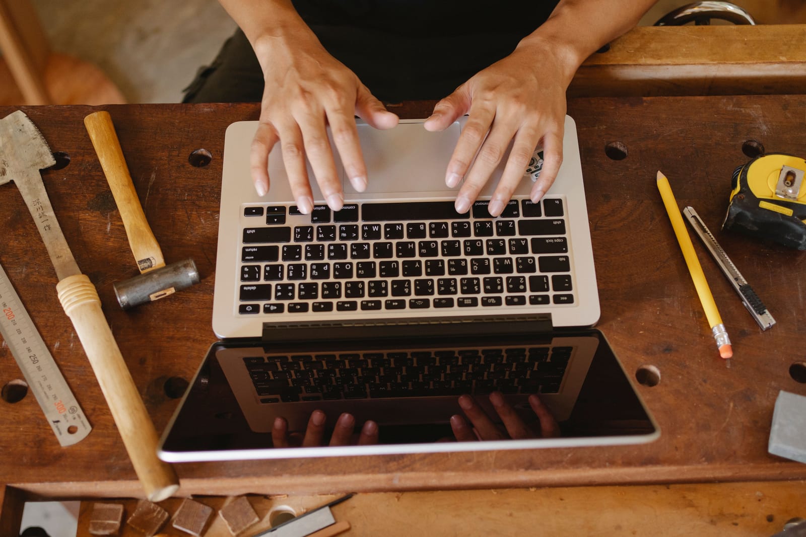 Hands typing on a laptop on a workbench, with a mallet, chisels and a tape measure beside it
